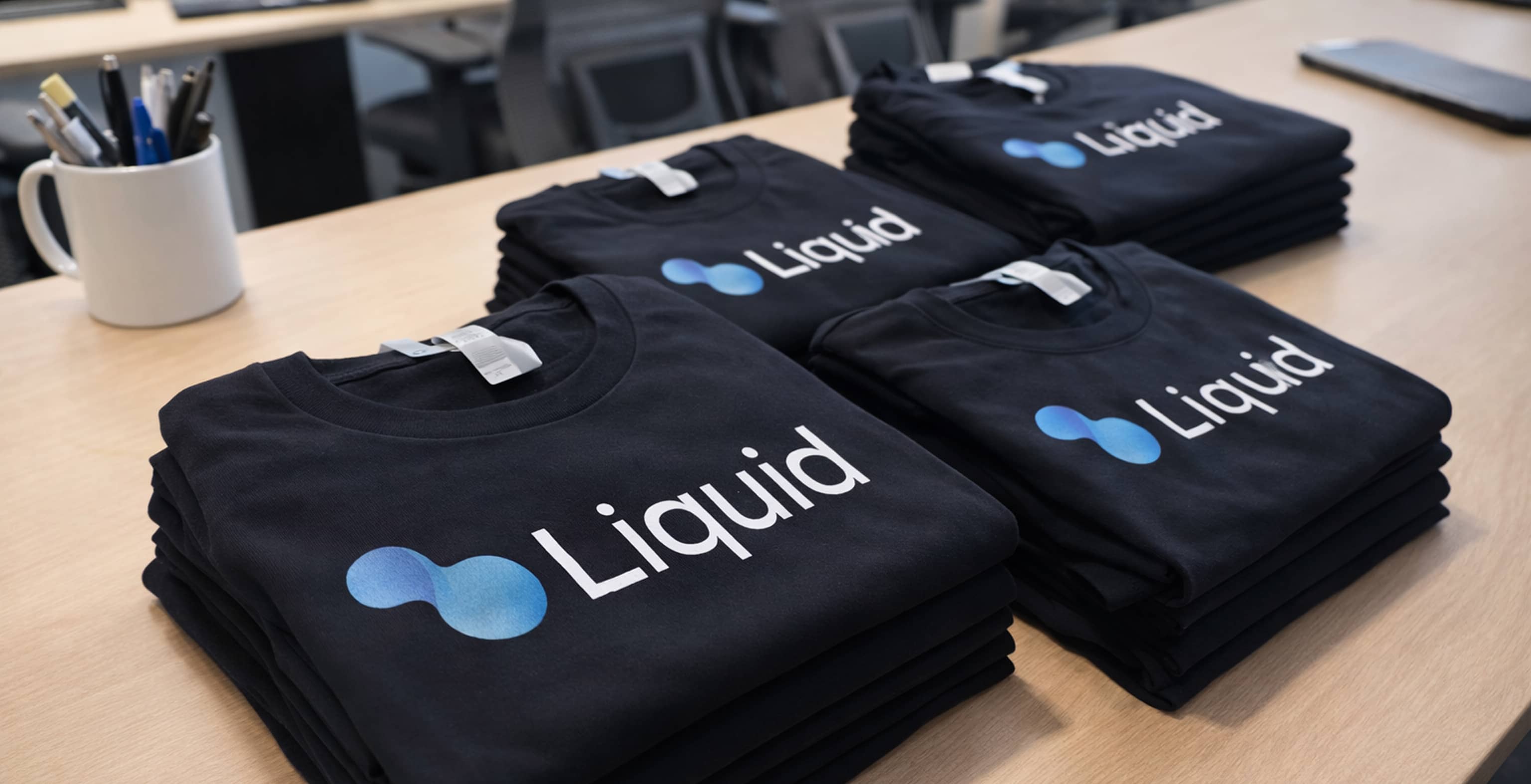 Stacks of folded navy t-shirts printed with the Liquid design system logo, laid out on an office table.